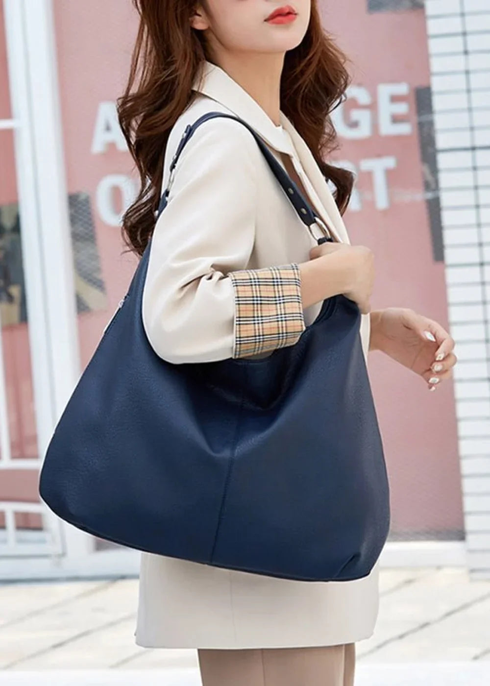 Navy blue slouchy tote bag with gold hardware worn by a woman in a cream blazer with plaid cuffs.