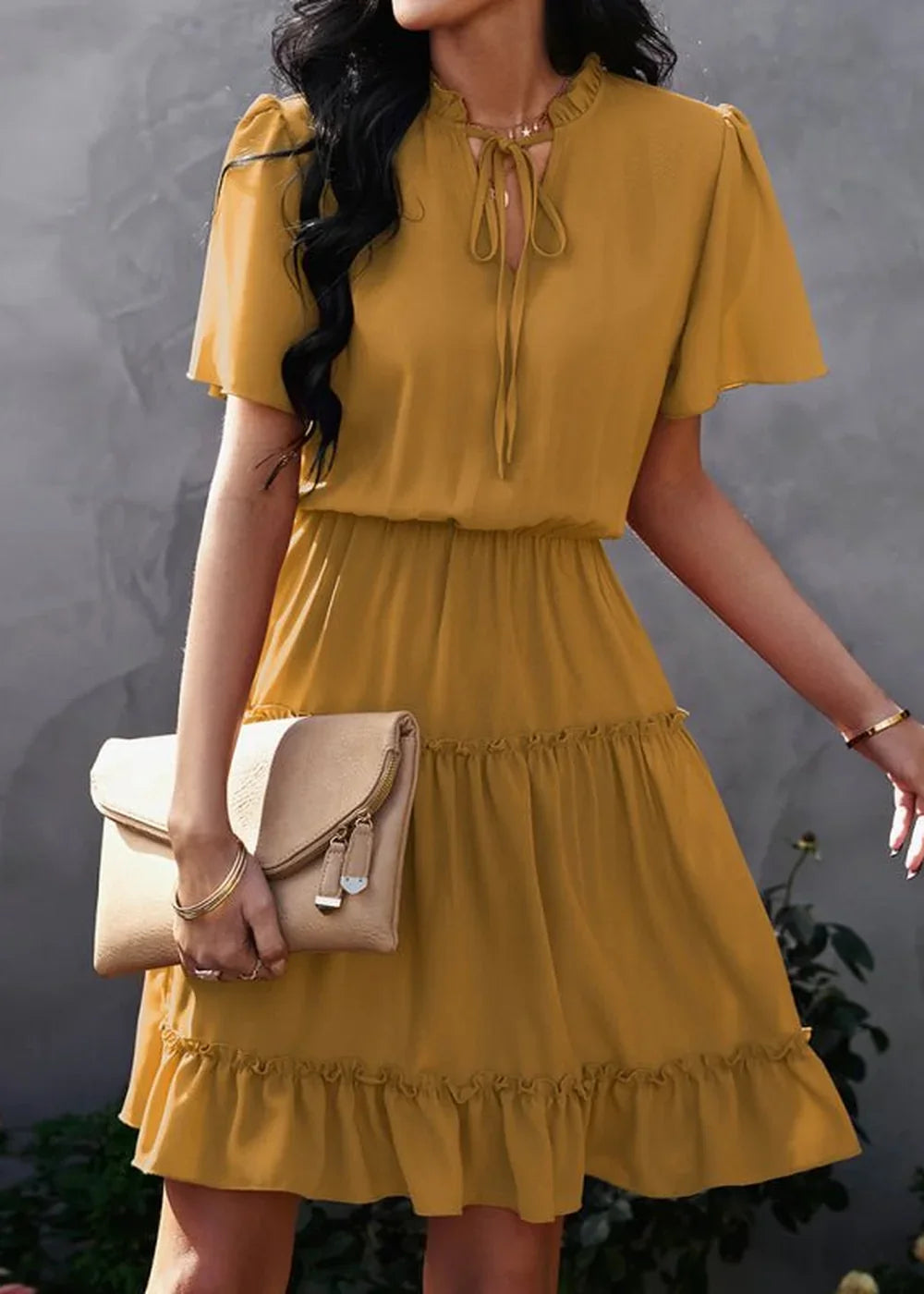 Mustard yellow tiered ruffle dress with tie-neck and flutter sleeves, held by a model with a clutch.