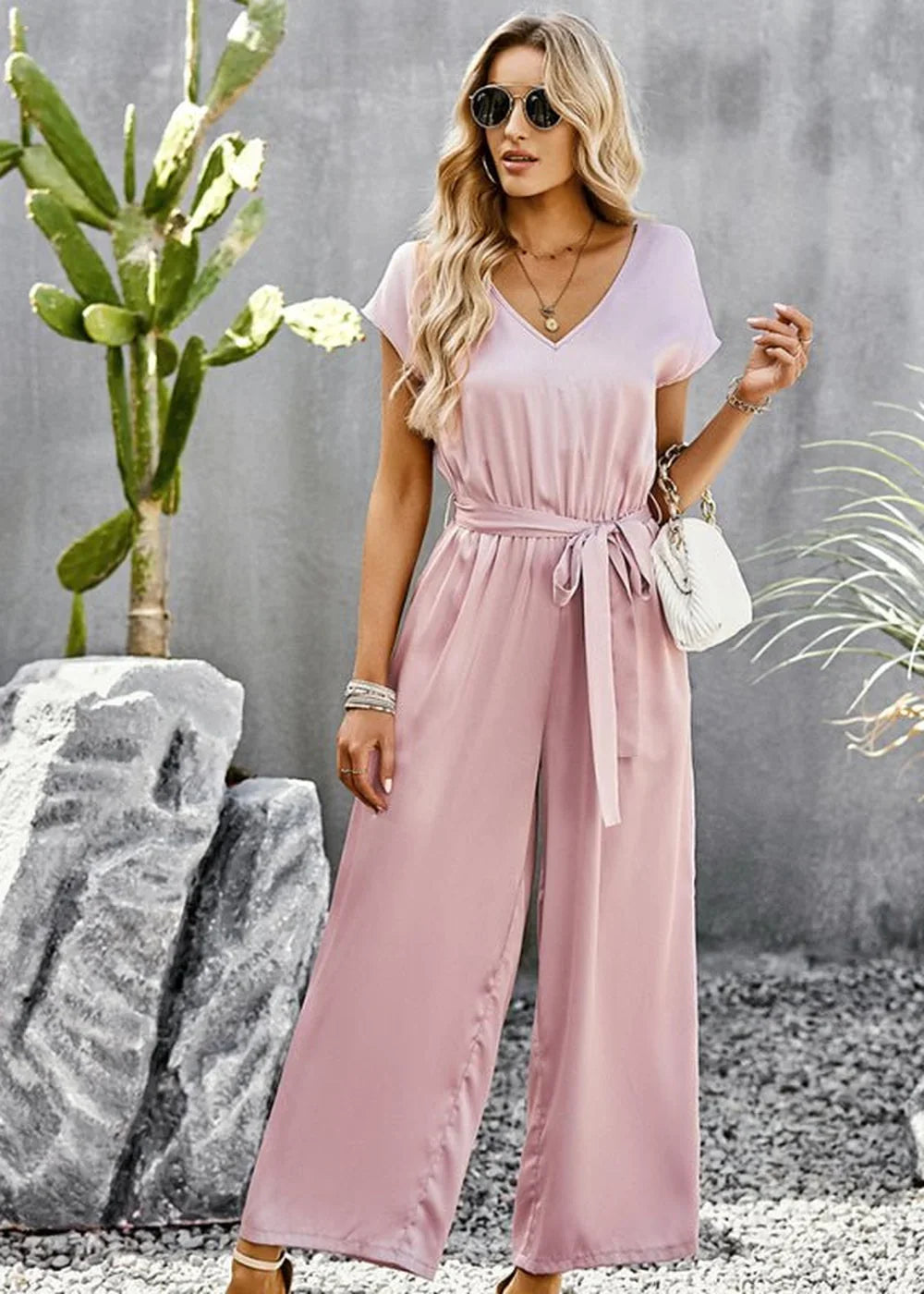Woman in a flowing pink satin jumpsuit with a V-neck and belted waist, standing outdoors.