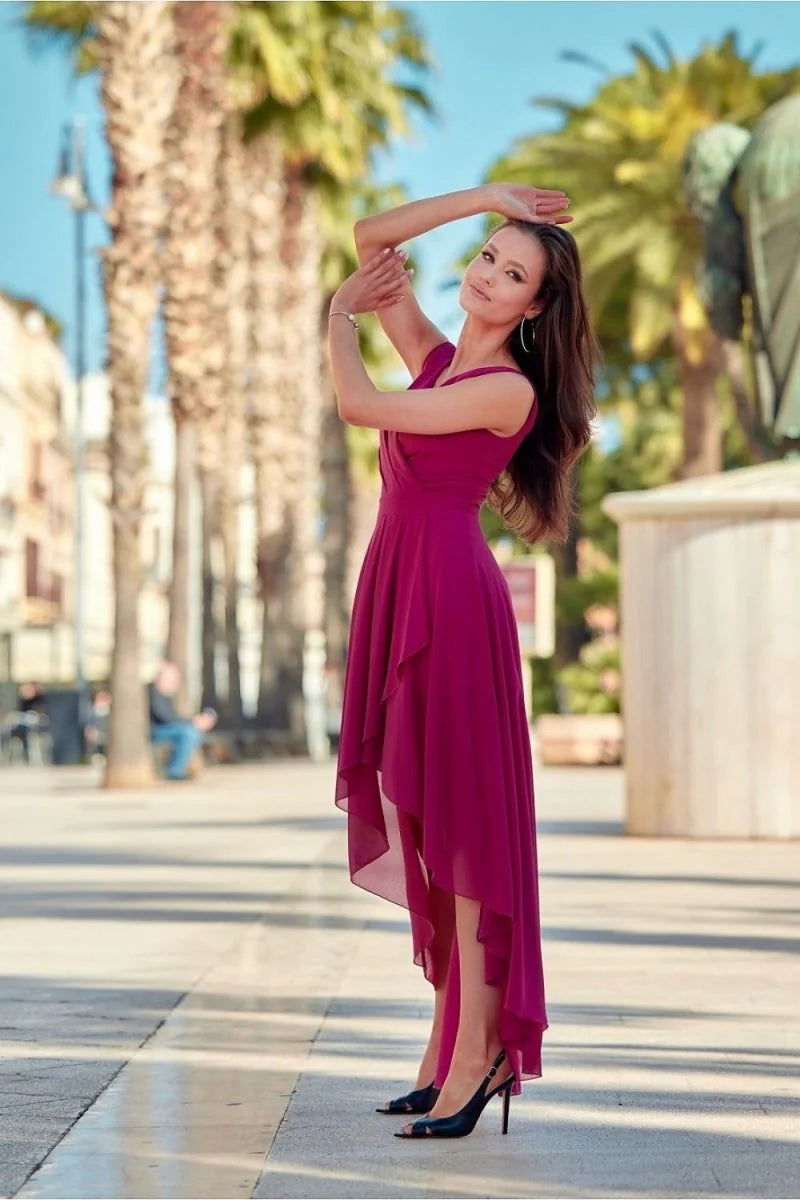 A woman in a flowing magenta dress with a high-low hem and black heels poses outdoors.