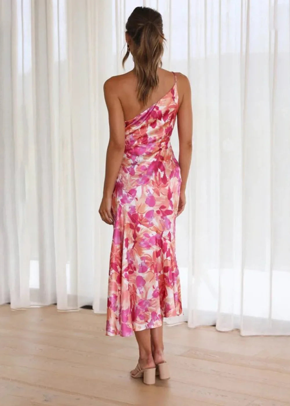 Back view of a woman in a flowy, one-shoulder floral print dress in shades of pink, orange, and white.