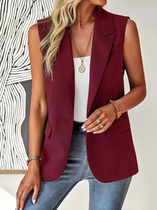 Sleeveless burgundy blazer with lapels, worn over a white top and jeans, accessorized with necklaces and rings.