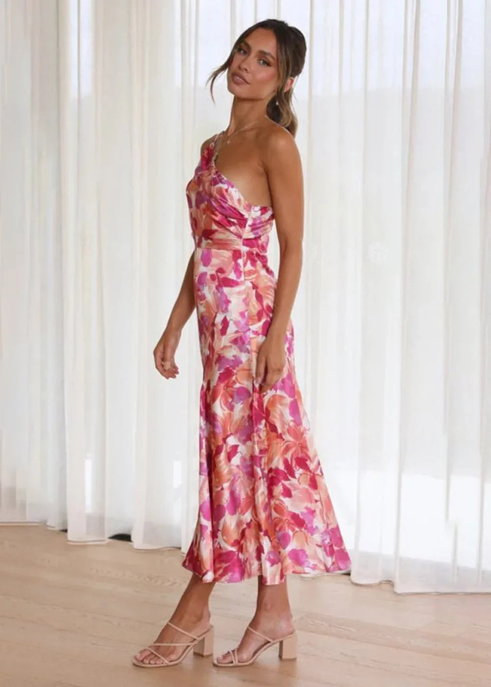 Full-length image of a woman wearing a pink and orange floral print one-shoulder midi dress and nude heeled sandals.