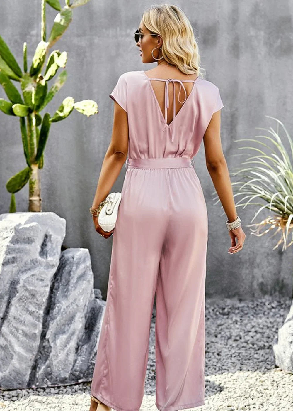 Blush pink satin jumpsuit with a tie-back V-neck and wide legs, shown from the back on a model.