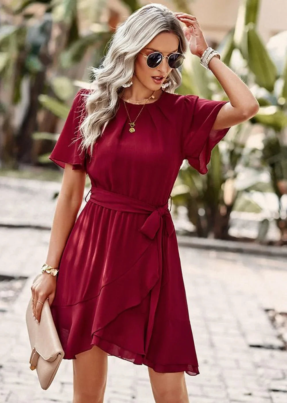 Woman in a flowing burgundy wrap dress with flutter sleeves and a tie waist, holding a light beige clutch.