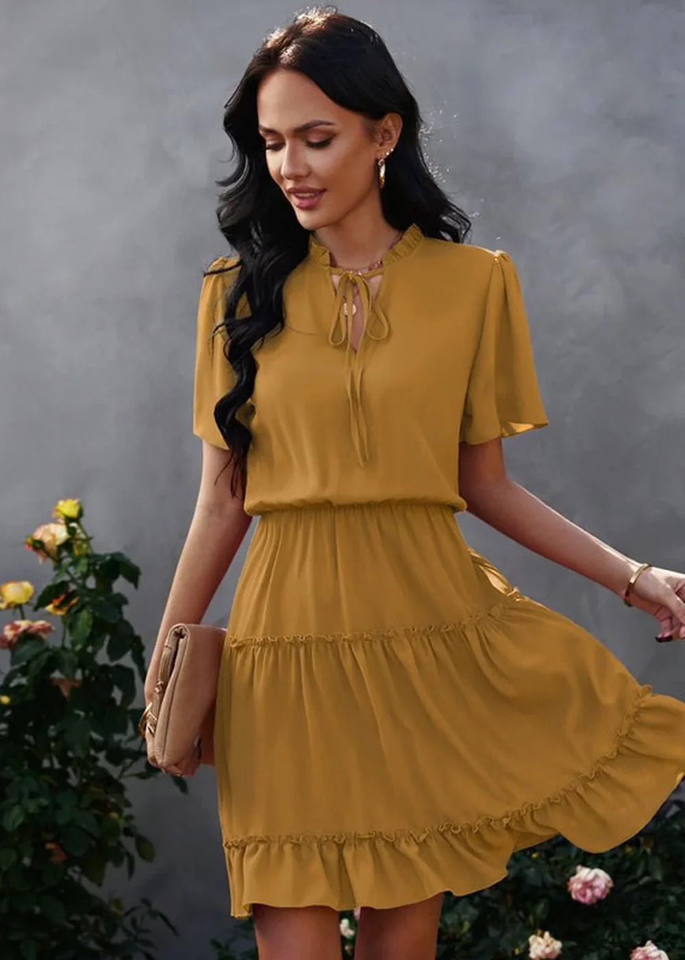 Mustard yellow tiered ruffle dress with tie-neck, shown on a model with a clutch.
