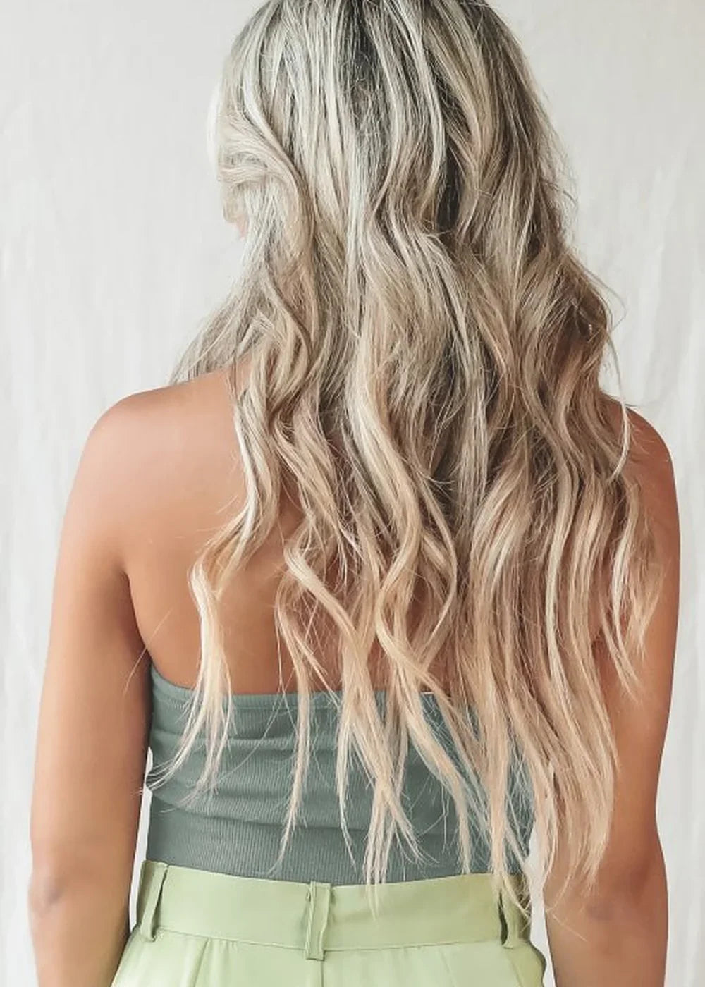 Back view of a woman with long, wavy blonde hair, wearing a green ribbed tube top and light green pants.
