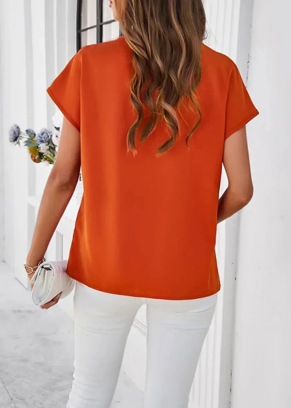 Back view of an orange polyester top with short sleeves, worn by a model with white pants and a white clutch.