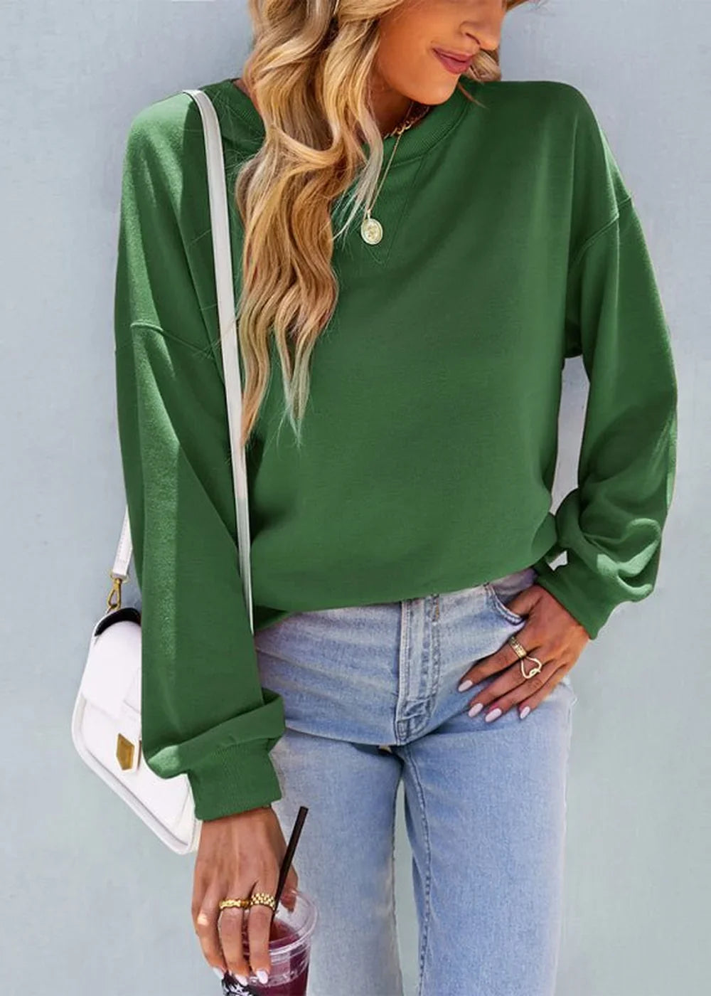Woman in loose-fit green crewneck sweatshirt and light wash jeans, holding a drink.