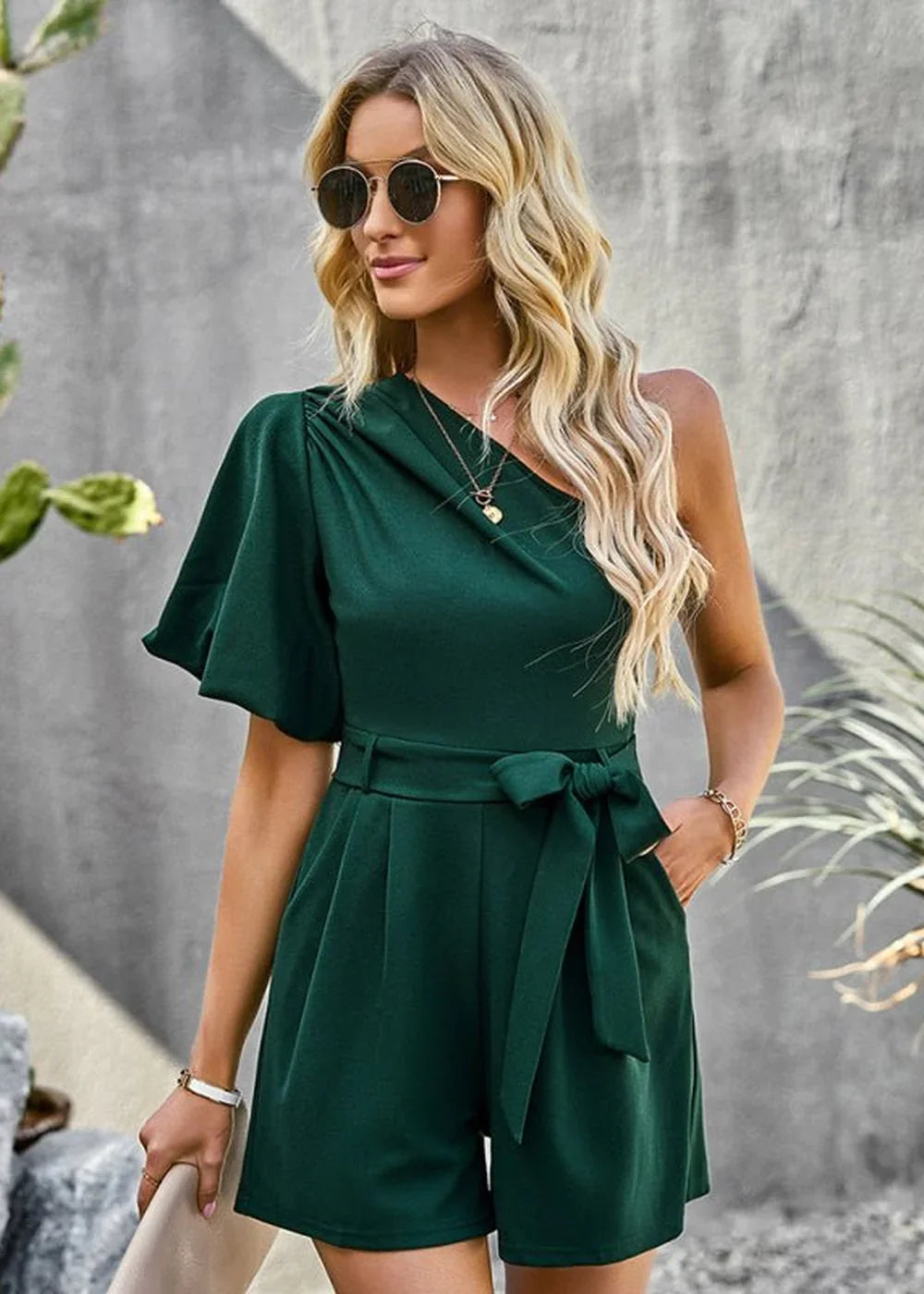 Woman in emerald green one-shoulder romper with tie waist and flutter sleeve, wearing sunglasses.