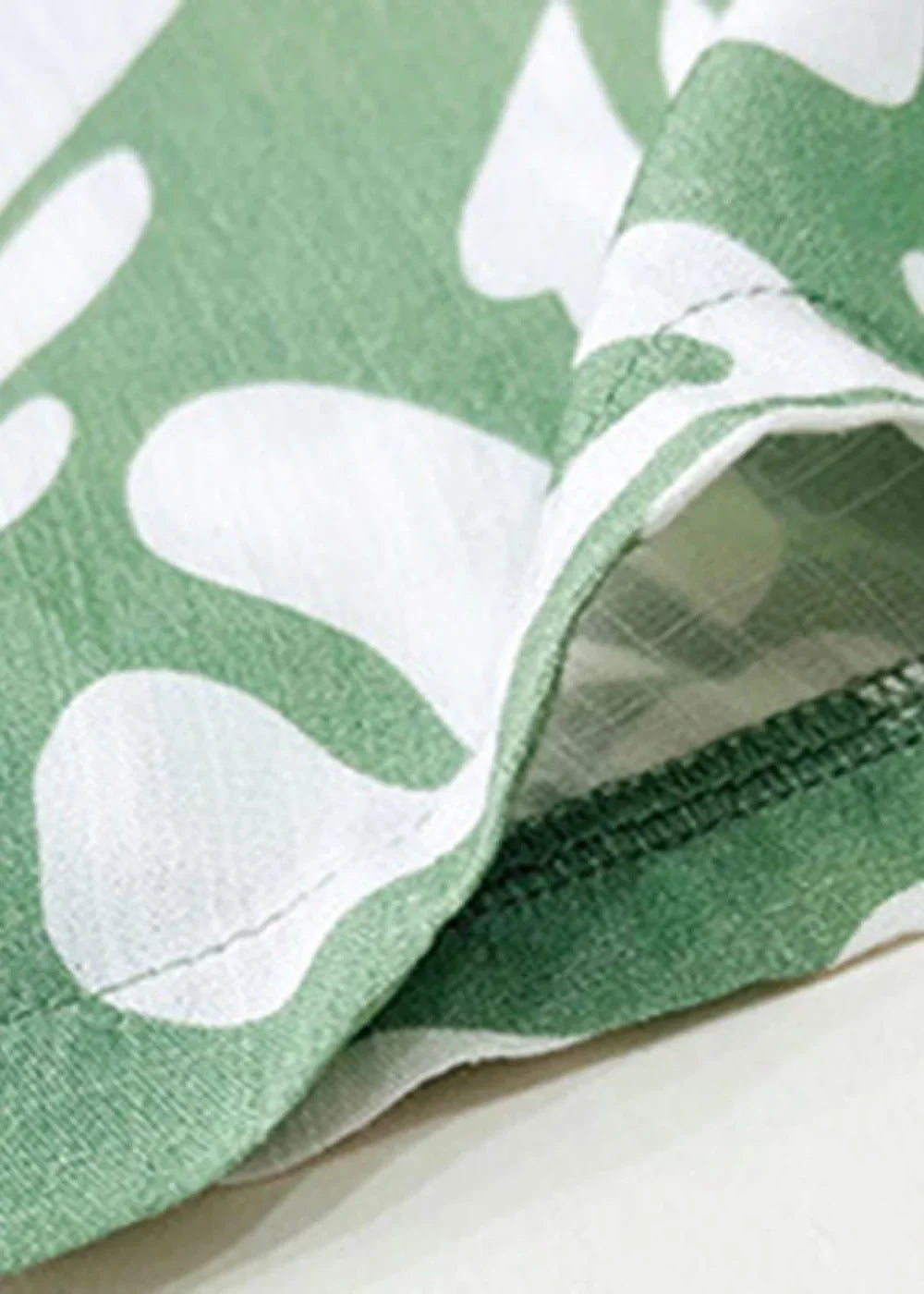 Close-up of green fabric with a white abstract leaf print and visible stitching on a hem.