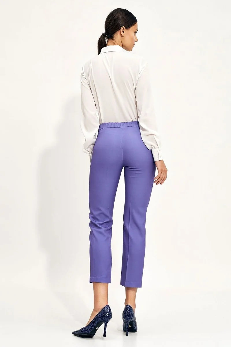 Back view of woman wearing high-waisted, cropped lavender trousers with a white blouse and dark blue snake print heels.