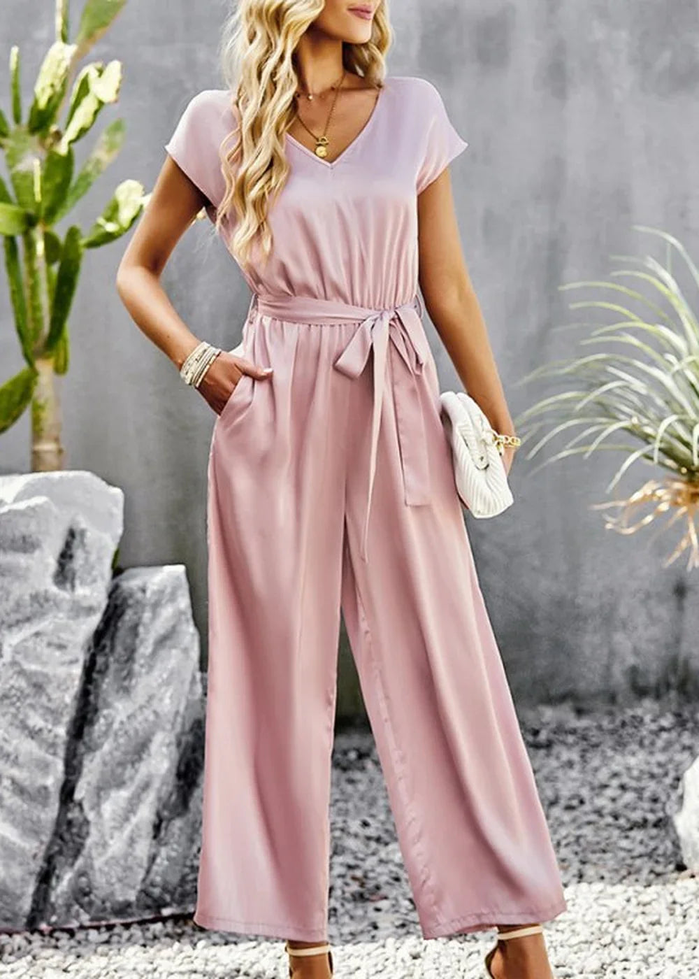 Blush pink satin jumpsuit with a V-neck, tie waist, and wide legs, worn by a model.