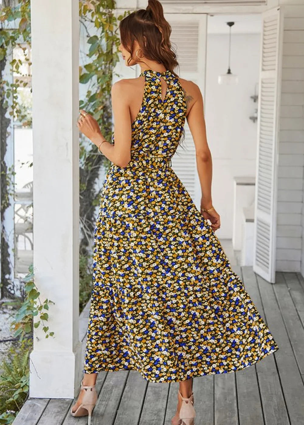 Back view of a woman in a yellow maxi dress with a black, blue, and white floral print and a halter neckline.