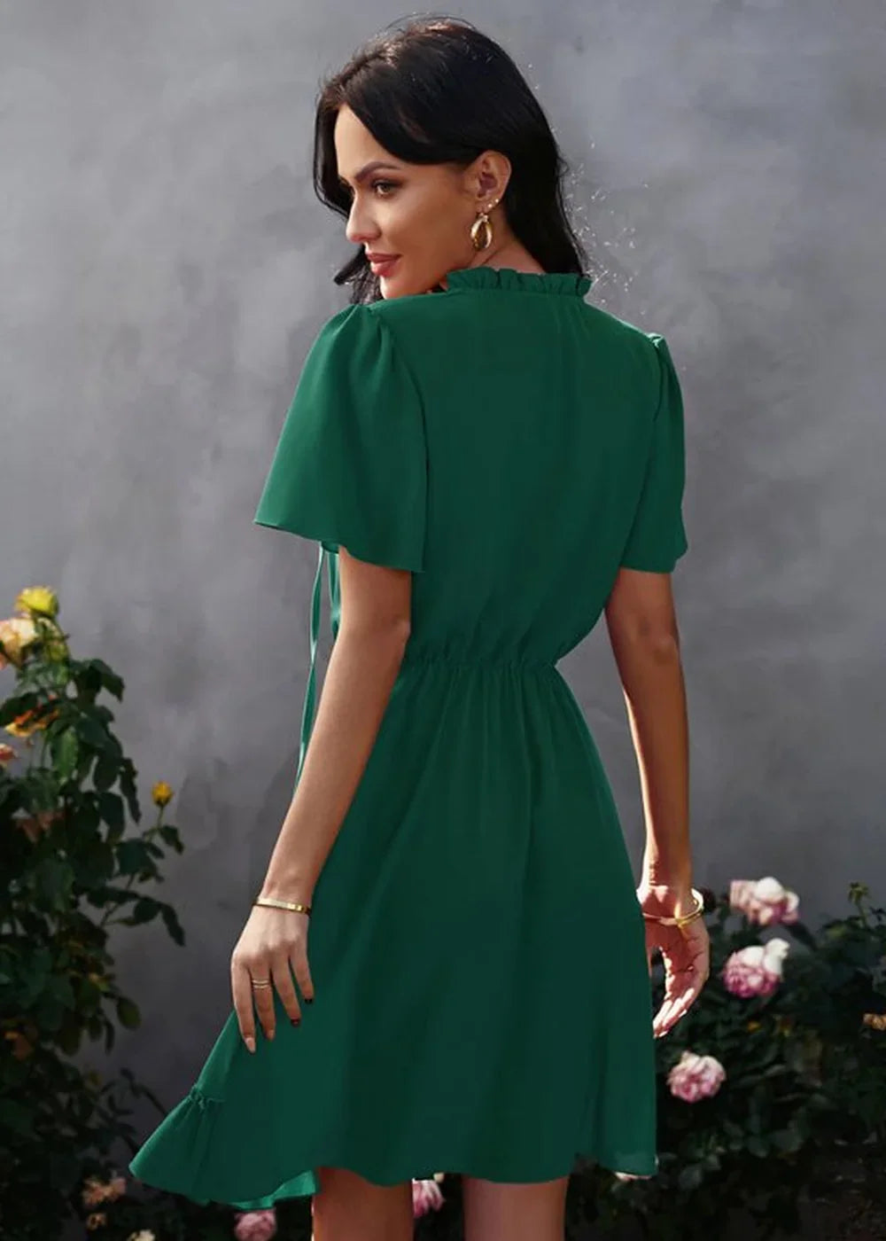 Woman in emerald green ruffled neckline dress with flutter sleeves, cinched waist, and ruffled hem, standing outdoors.