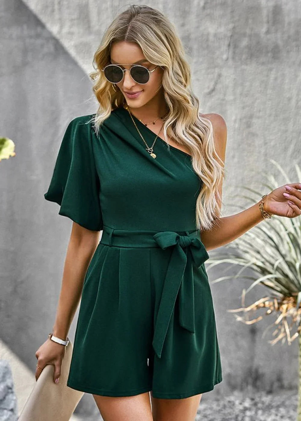 Woman in a dark green one-shoulder romper with a belted waist and a flowing sleeve.
