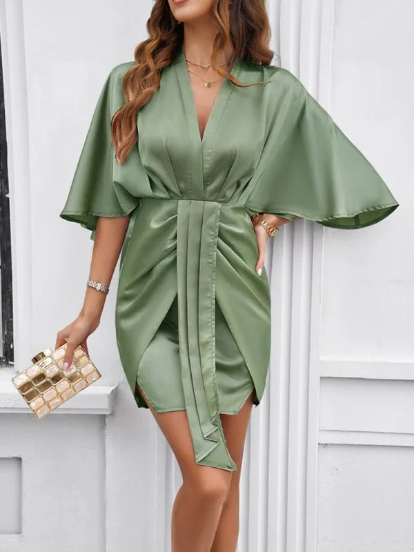 Sage green satin wrap dress with flutter sleeves and ruched detailing, held with a gold clutch.