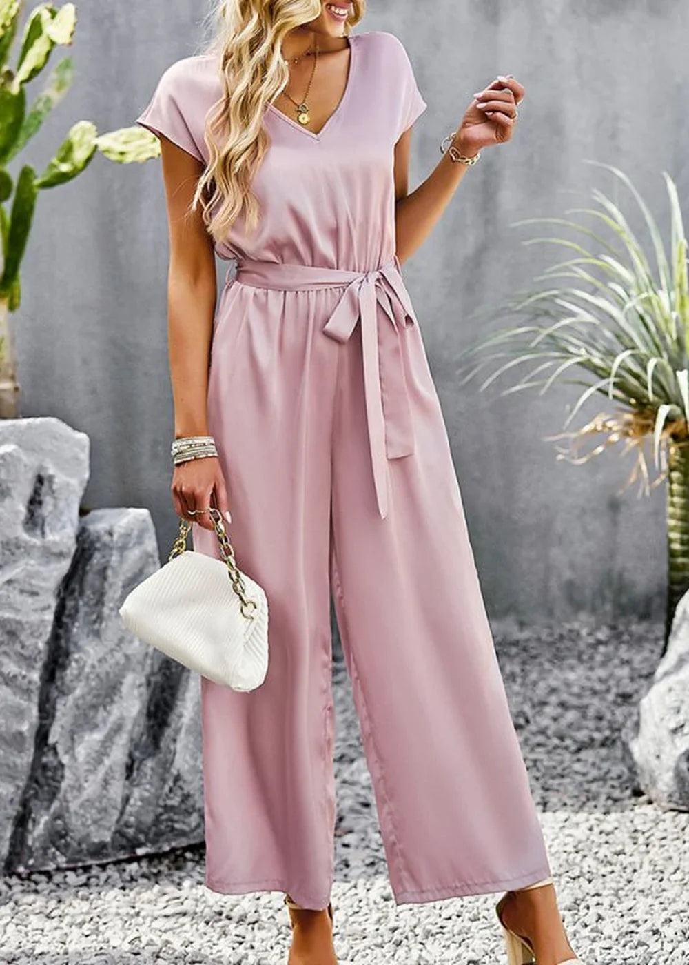 Pink satin jumpsuit with V-neck, tie waist, and wide legs, held by a model.