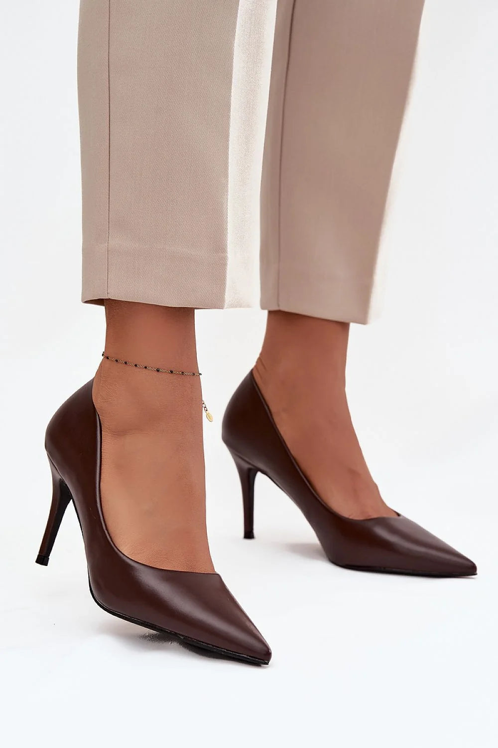 Brown pointed-toe pumps with stiletto heels and an ankle bracelet with a gold charm.