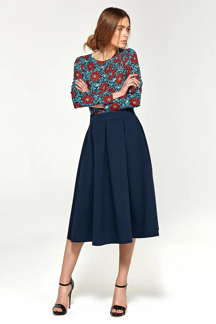 Woman in a navy blue pleated midi skirt and a long-sleeved top with a red and blue floral pattern, wearing black high heels.