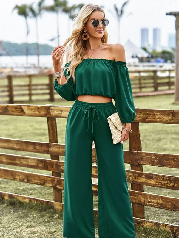 Woman in a forest green off-the-shoulder crop top and wide-leg pants, holding a woven clutch.