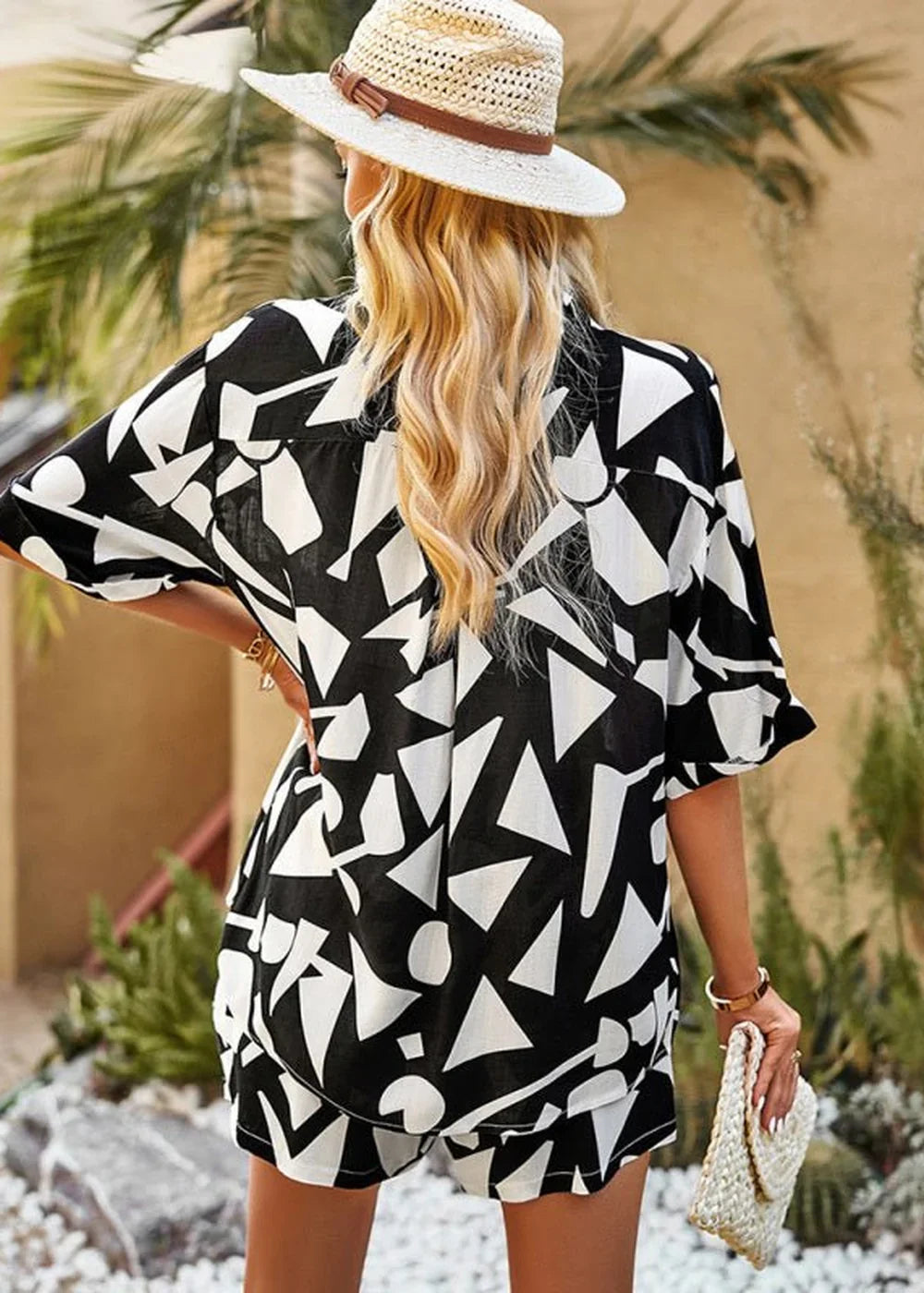 Woman in a black and white geometric print short suit and straw hat, holding a clutch.