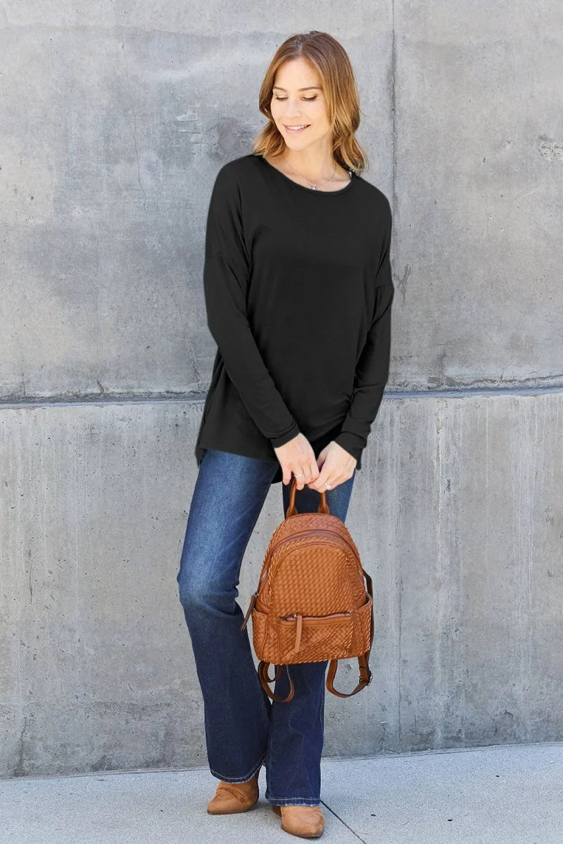 Woman in black long-sleeve top and flared jeans holding a woven brown backpack.