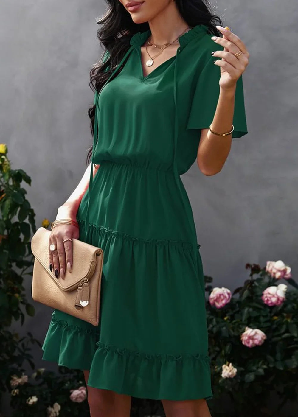 Emerald green tiered ruffle dress with a V-neck and tie detail, held by a model with a tan clutch.