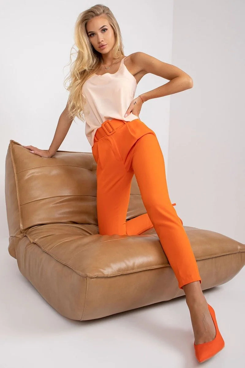 Orange high-waisted pants with a matching belt, worn by a model on a tan chair.