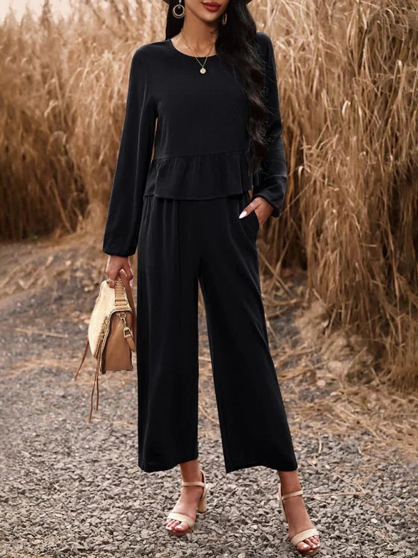 Black wide-leg jumpsuit with ruffle detail at the waist, worn by a model with tan heeled sandals and a straw bag.