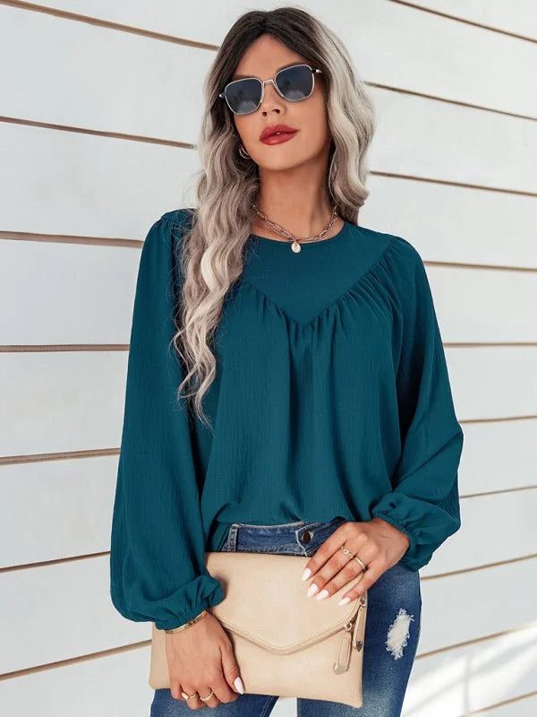 Teal long-sleeved blouse with gathered neckline and billowy sleeves, worn by a model holding a beige clutch.