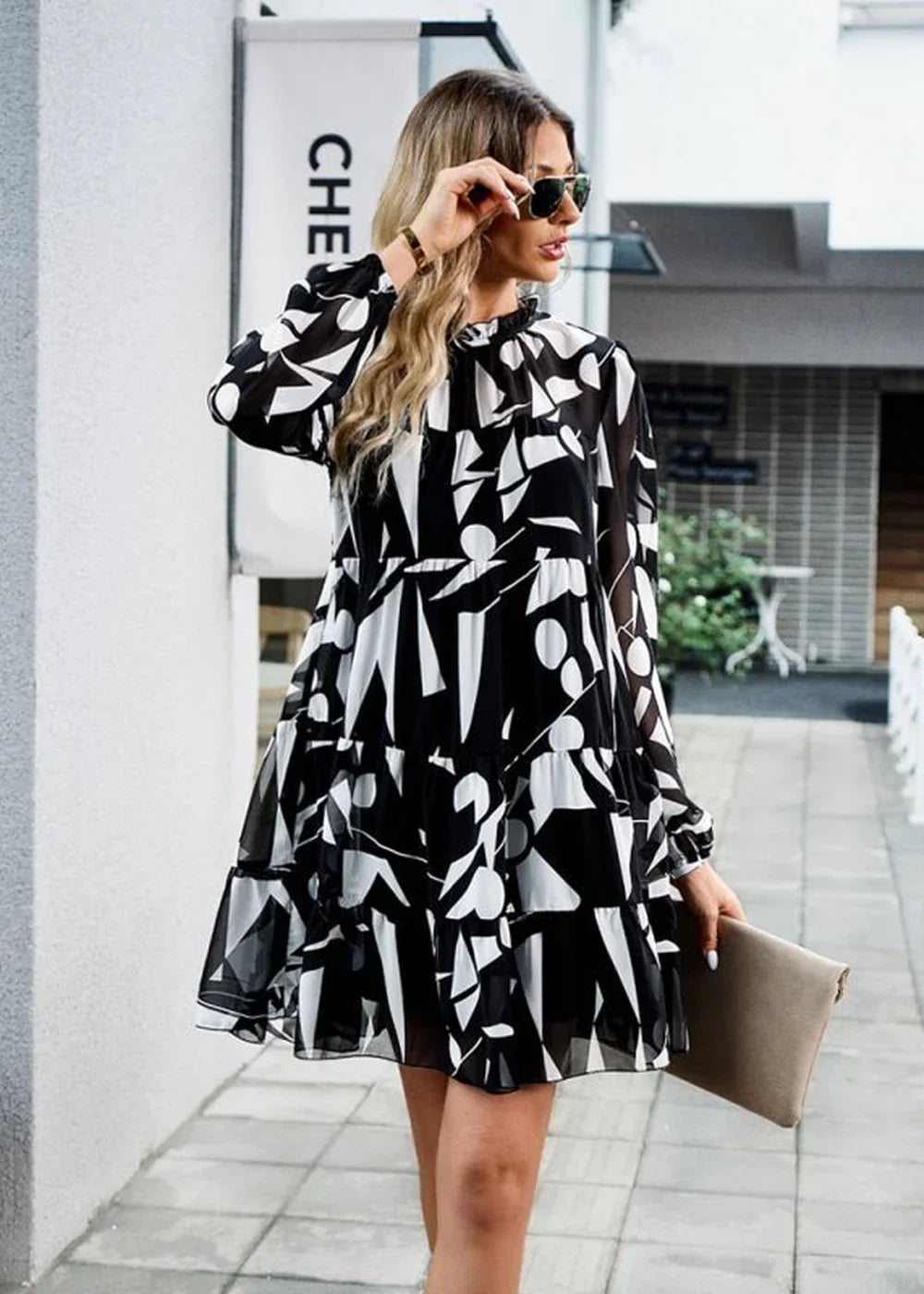 Black and white abstract print tiered mini dress with long sleeves and a mock neck, held by a woman on a sidewalk.