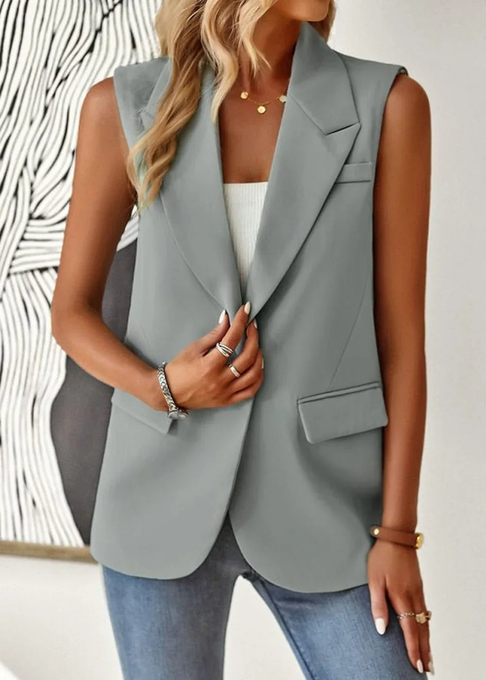 Woman wearing a light gray sleeveless blazer over a white top, with blue jeans and jewelry.