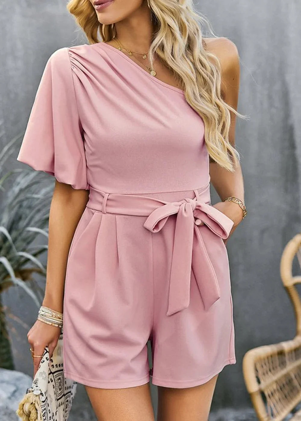 Blush pink one-shoulder romper with flutter sleeves, self-tie belt, and pleated shorts.