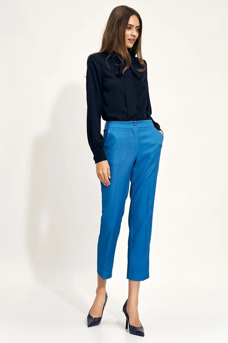 Full-length view of a woman wearing bright blue capri pants and a black bow-neck blouse.