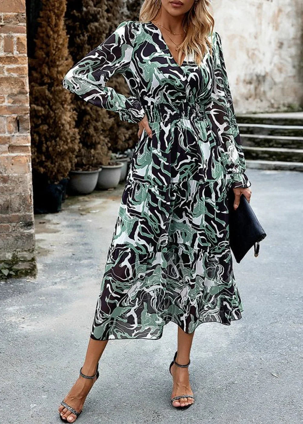 Woman in a green, black, and white abstract print tiered maxi dress with a V-neck and long sleeves, holding a black clutch.