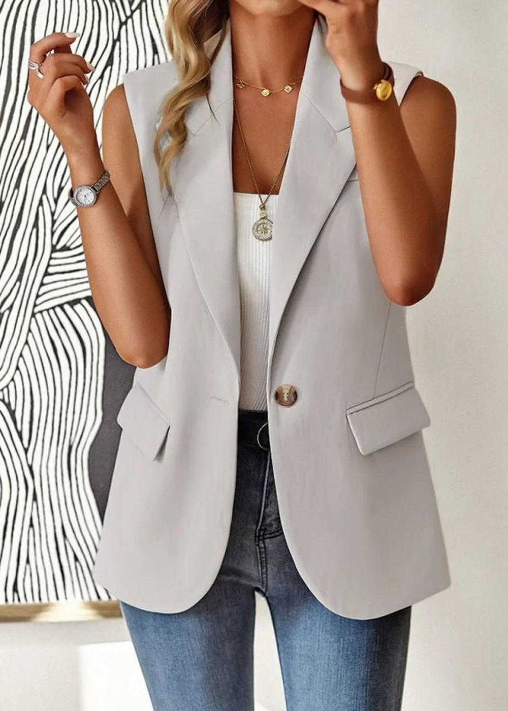 Light grey sleeveless blazer with a single button closure and front pockets, worn by a model over a white top and blue jeans.
