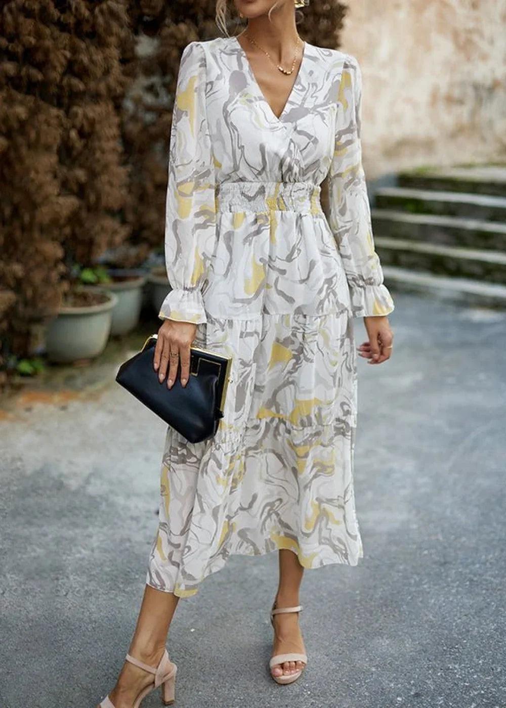 Woman in a white, yellow, and gray abstract print tiered midi dress with smocked waist and ruffled cuffs, holding a black ...