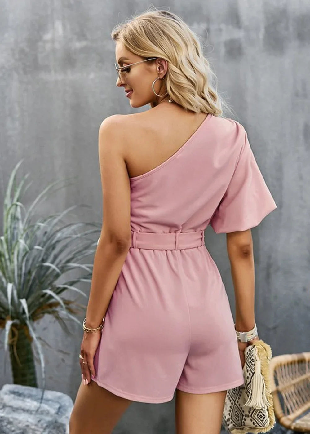 Pink one-shoulder romper with belted waist and ruffled sleeve detail.