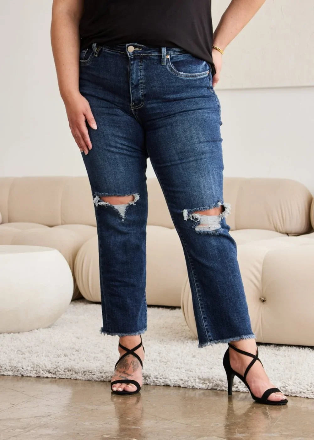 Dark wash distressed denim jeans with frayed hem and knee rips, worn by a model with black heels.
