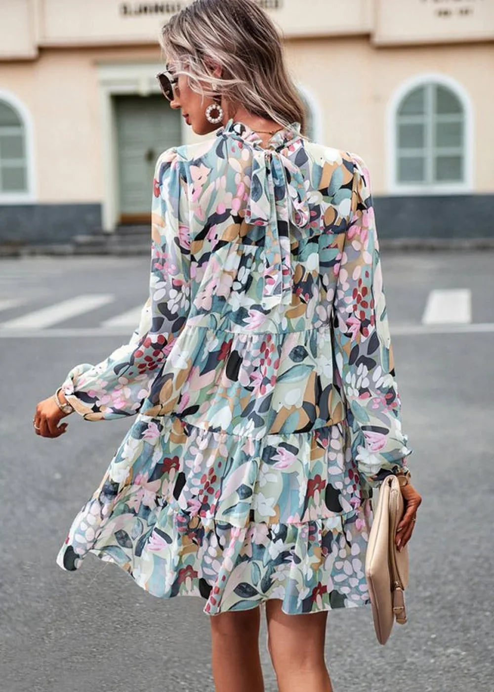 Flowy floral print dress with a tiered skirt and a bow tie at the neck in pastel colors.