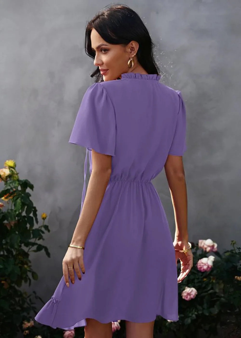 Back view of a woman wearing a flowy purple polyester dress with ruffled neckline and short sleeves.