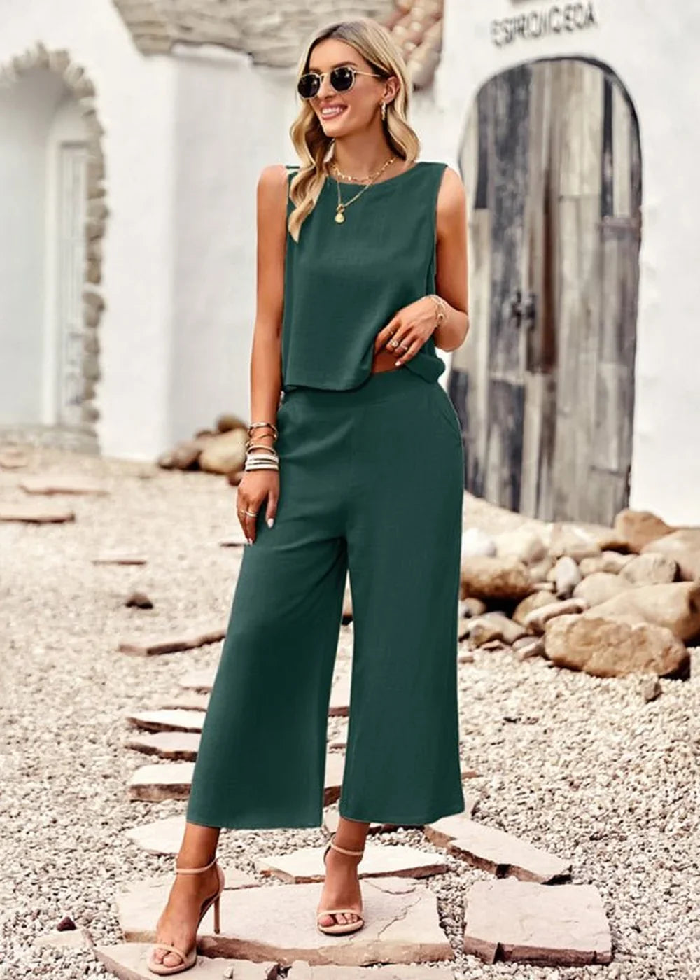 Woman in a dark green sleeveless crop top and wide-leg pants, wearing sandals and sunglasses.