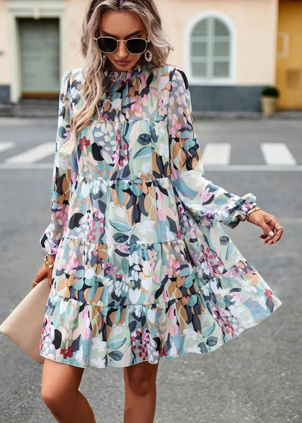 Woman in flowy, tiered floral print mini dress with long sleeves and a high neckline, holding a clutch.