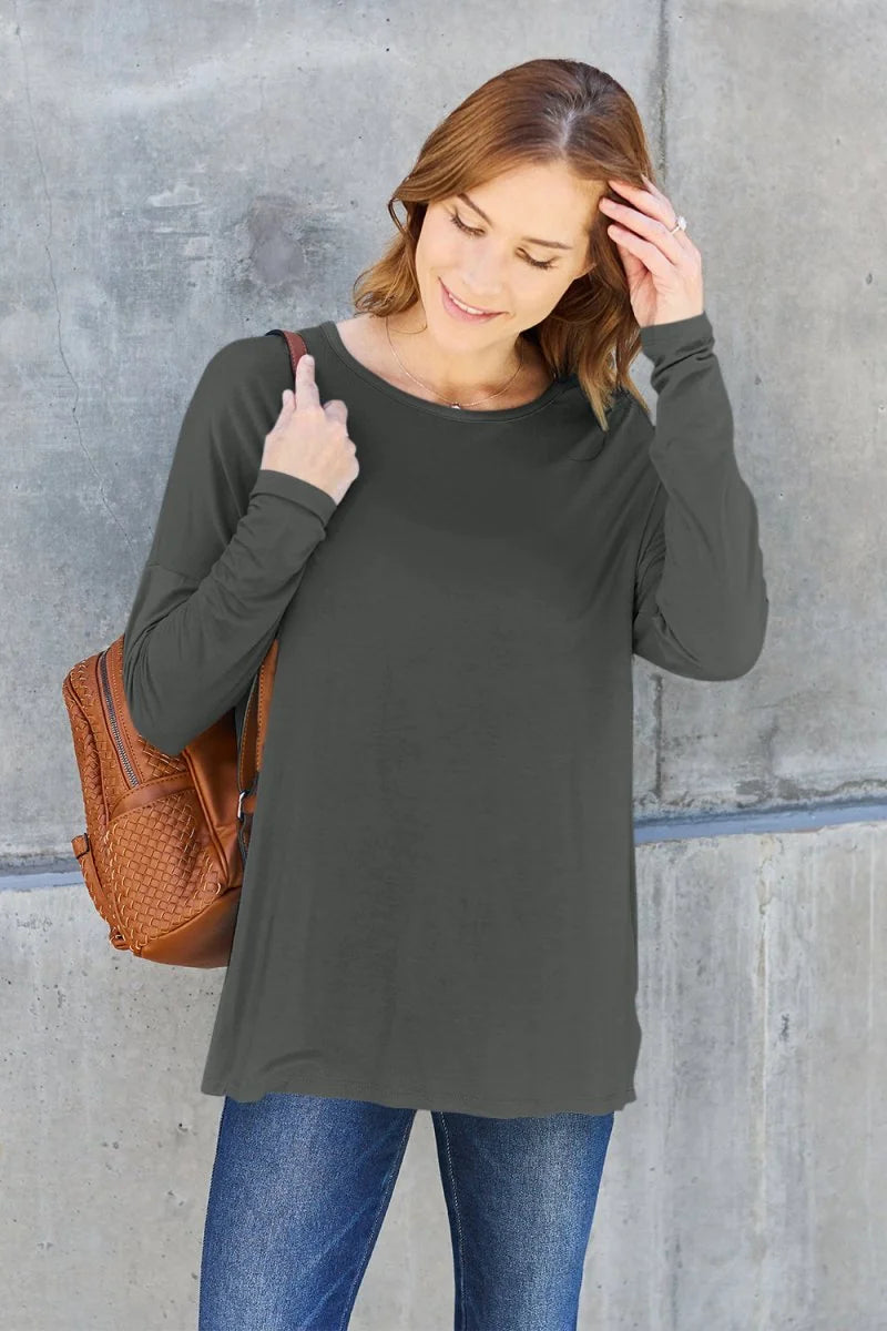 A woman wearing a charcoal long-sleeve top and blue jeans, with a brown woven backpack on her shoulder.
