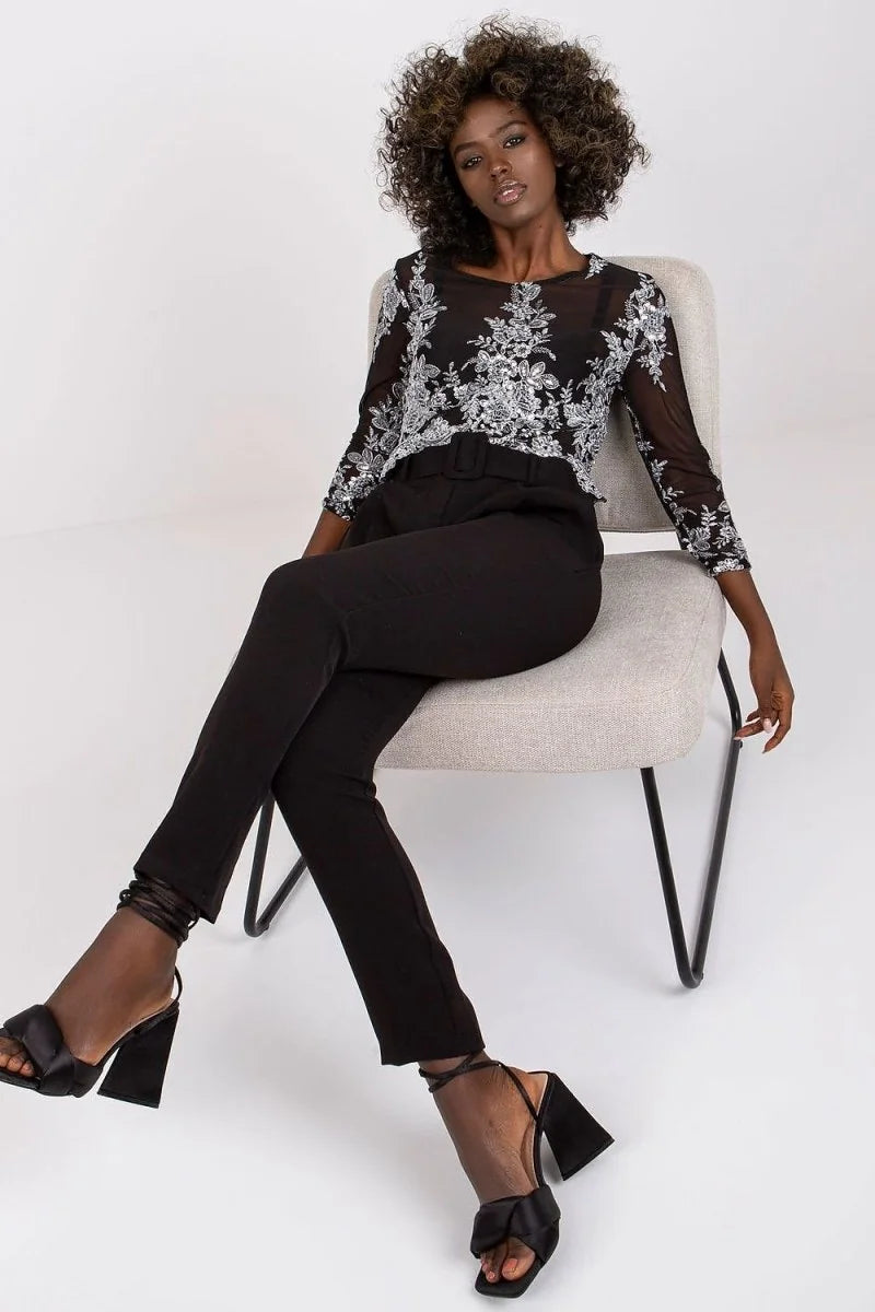 Black pants with a belt and sheer, embroidered top with long sleeves worn by a model.