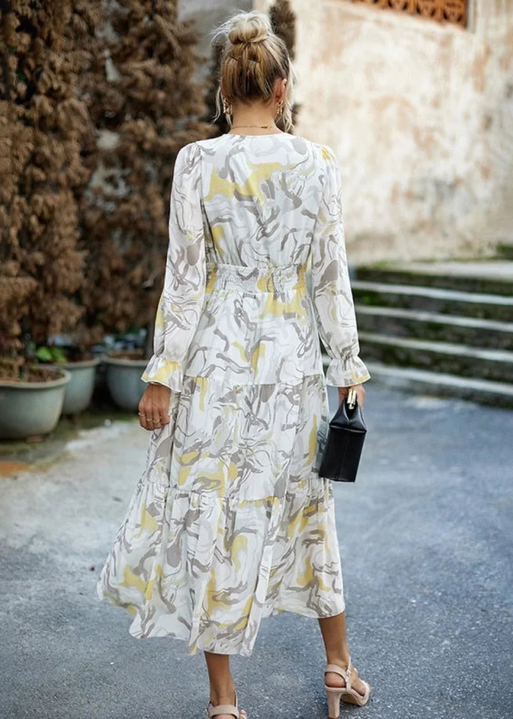 Woman from behind in a flowing white dress with a yellow and grey abstract print and tiered skirt.