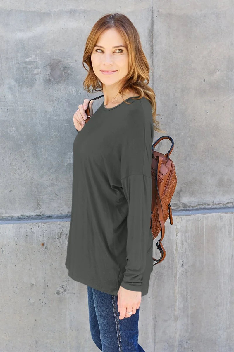 Model in a charcoal grey long-sleeve top and blue jeans, carrying a brown woven backpack.