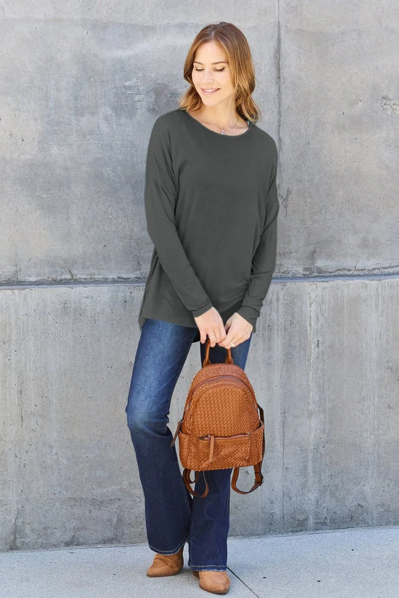 Woman in dark grey long-sleeve top, blue flare jeans, and tan shoes holding a woven brown backpack.