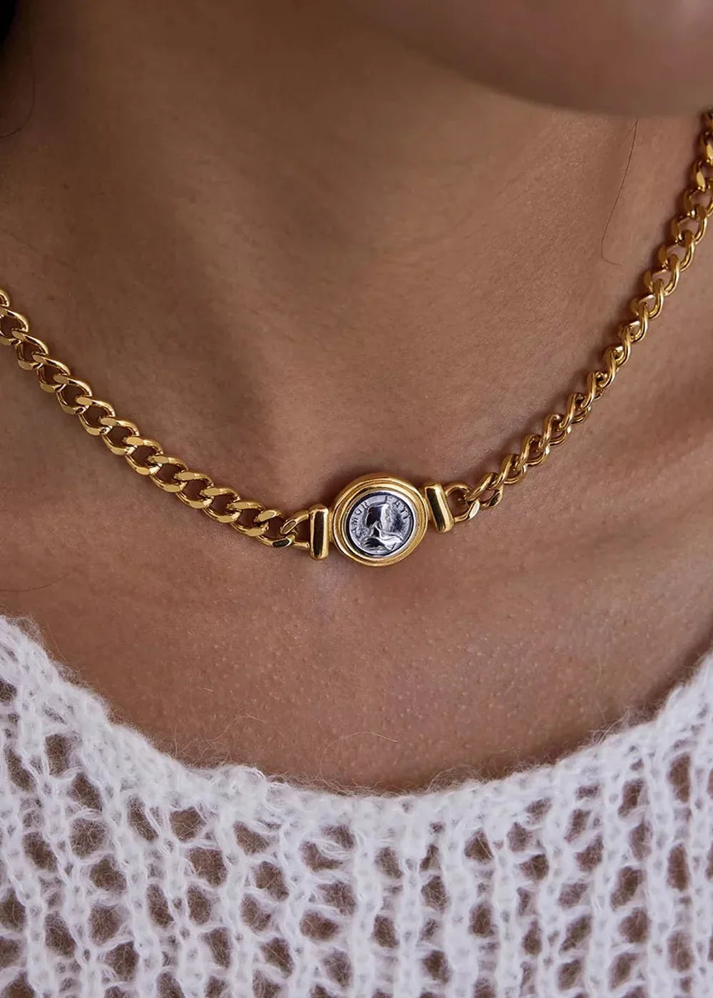 Gold chain necklace with a circular coin pendant, worn on a person's neck over a white knit top.
